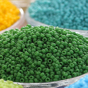 Close-up of small round craft beads in multiple colors, with a prominent mound of green beads in the foreground and bowls of yellow, turquoise, light green, and blue beads blurred in the background.